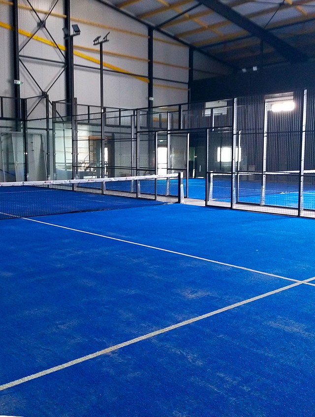 Terrain de padel indoor aux sols bleus, filet et parois vitrées dans le complexe Indoor Padel Loudéac